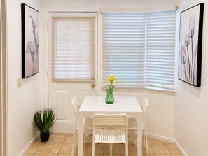 Una mesa de comedor blanca con cuatro sillas, un jarrón verde con flores amarillas y dos cuadros en las paredes.