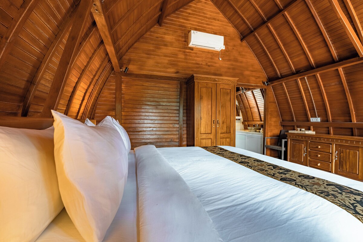 A bedroom with a wooden ceiling, a bed with white linens, a wooden dresser, and a small kitchenette area.