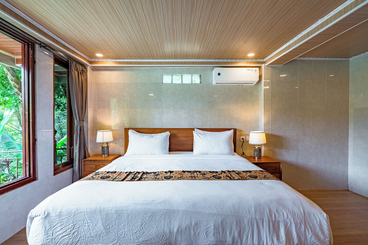 A hotel room with a large bed, two bedside tables with lamps, a ceiling with wooden paneling, and a window with curtains.