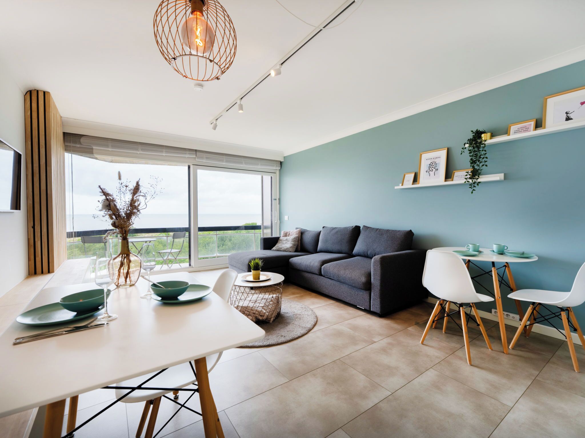 A modern living room with a sofa, dining table, and chairs, a balcony with a view, and a ceiling light fixture.