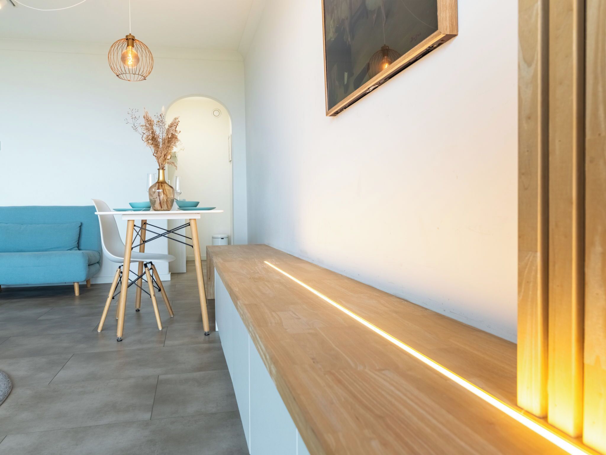 A modern living room with a blue sofa, a dining table with chairs, a vase with dried flowers, and a pendant light.