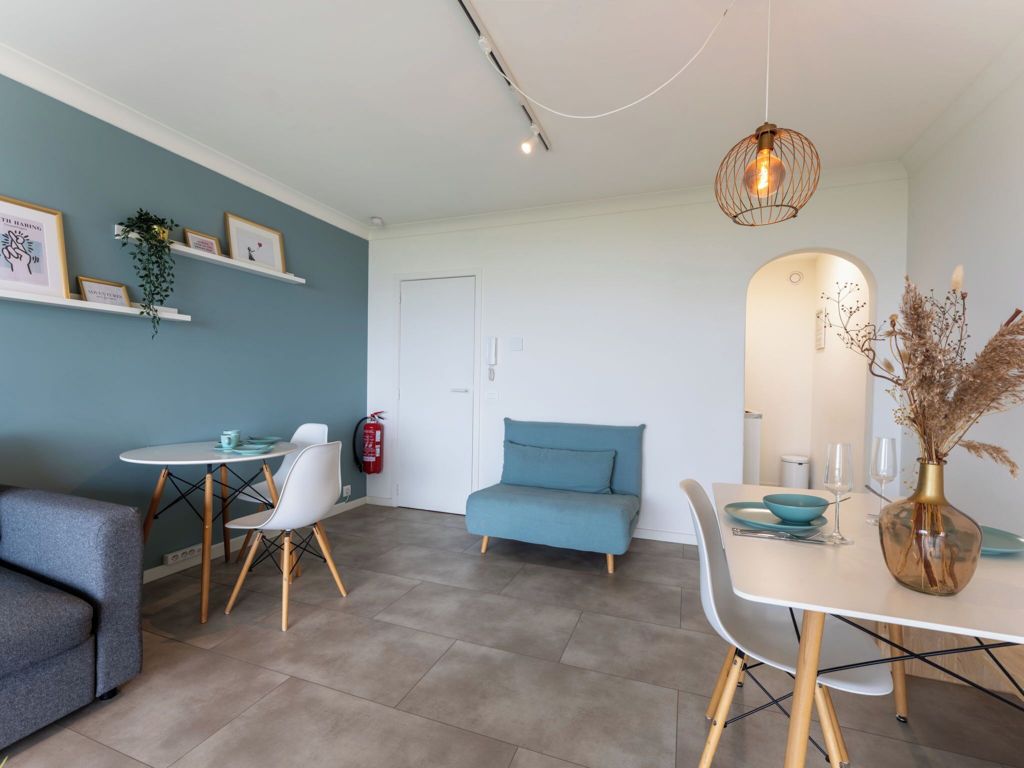 A modern living room with a sofa, a dining table with chairs, a blue armchair, a small table with a bowl and wine glasses, and a vase with dried plants.