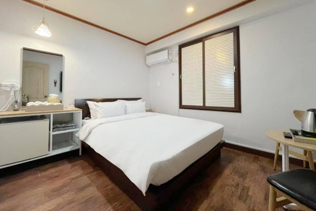 A hotel room with a bed, bedside table, mirror, and a window with blinds.