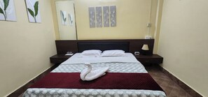 A neatly made bed with a swan-shaped pillow, a bedside table with a lamp, and artwork on the wall. - Hotel Roy's (Port Blair)