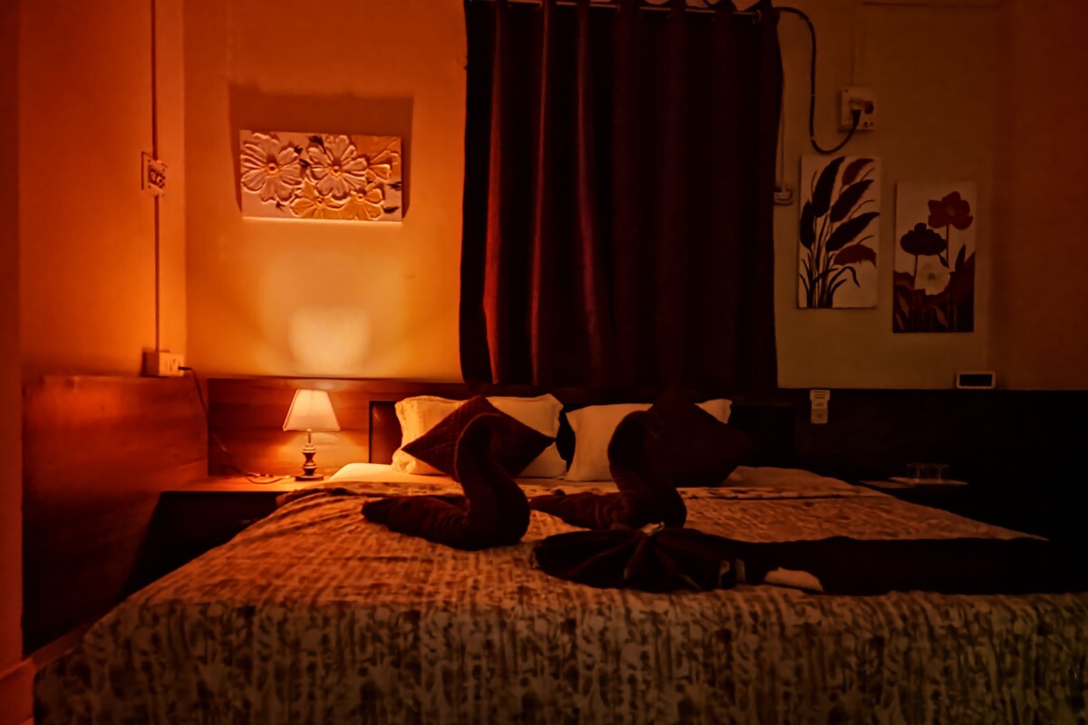 A dimly lit hotel room with a bed, a nightstand, a lamp, and a wall-mounted artwork.
