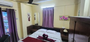 Ein Hotelzimmer mit Bett, Deckenventilator, Fenster mit VorhÀngen, Nachttisch mit Lampe und einem an der Wand befestigten Spiegel.
