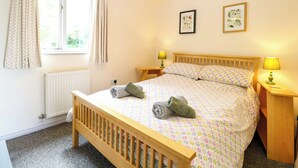 Quarto com cama de madeira, duas luminárias de mesa, uma janela com cortinas, um radiador e duas molduras com quadros na parede.