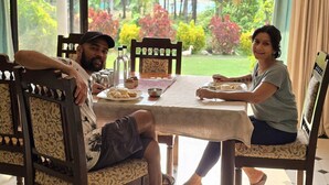 Un comedor con una mesa lista para dos, sillas y vista a un jardín con diversas plantas y césped.