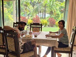 A dining area with a table set for two, chairs, and a view of a garden with various plants and a lawn.