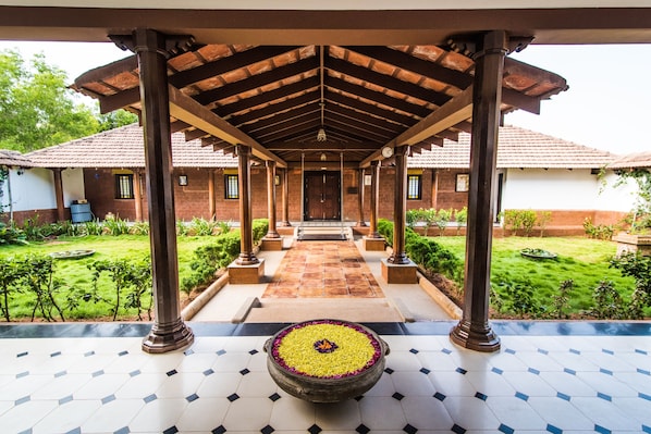 A covered walkway with a tiled floor leading to a building entrance, surrounded by greenery.