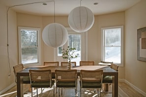 Een eetkamer met een grote tafel, zes stoelen, twee hangende papieren lampions en een vaas met groen.
