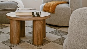 Uma sala de estar com um sofá cinzento, uma mesa de centro redonda de madeira e uma grande janela com cortinas.