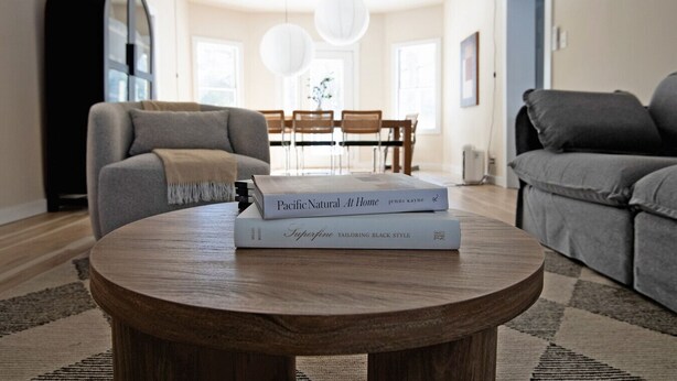 Uma sala de estar com uma mesa de centro redonda de madeira, dois livros, um sofá cinza, um armário preto e uma área de jantar ao fundo.