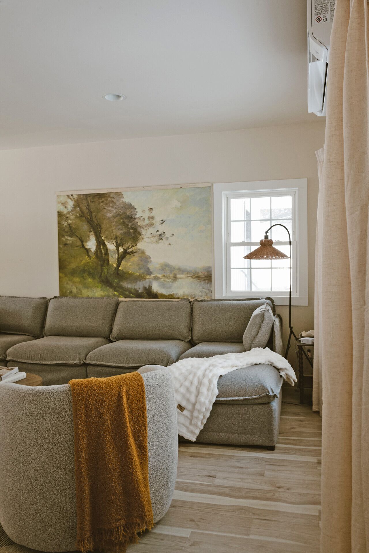 A living room with a sofa, a round ottoman, a painting, a window, and a lamp.