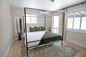 Una cama con dosel con una colcha blanca y verde, una mesita de noche pequeña, un espejo y una ventana con cortinas.
