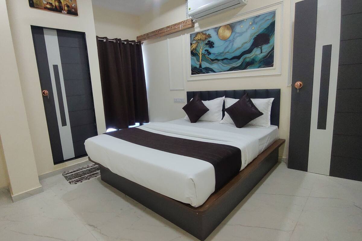 A neatly made bed with a headboard, a dark-colored wardrobe, a painting on the wall, and a window with curtains.