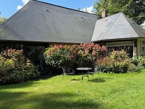 Une maison avec un toit en pente, un patio couvert avec une table et des chaises, et un jardin bien entretenu agrémenté d’arbustes en fleurs.