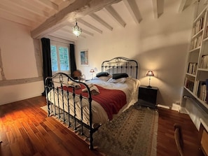 Une chambre à coucher avec un lit, des tables de chevet, une bibliothèque, une fenêtre avec des rideaux et un plancher en bois.