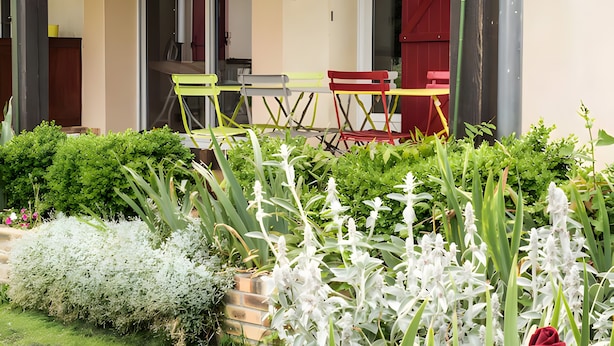 Un espacio exterior cubierto con una mesa y sillas, una puerta que da al interior, y un jardín con diversas plantas y flores.