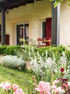 Et overdækket udendørs opholdsområde med et bord og stole, en dør ind til bygningen og en have med forskellige planter og blomster.