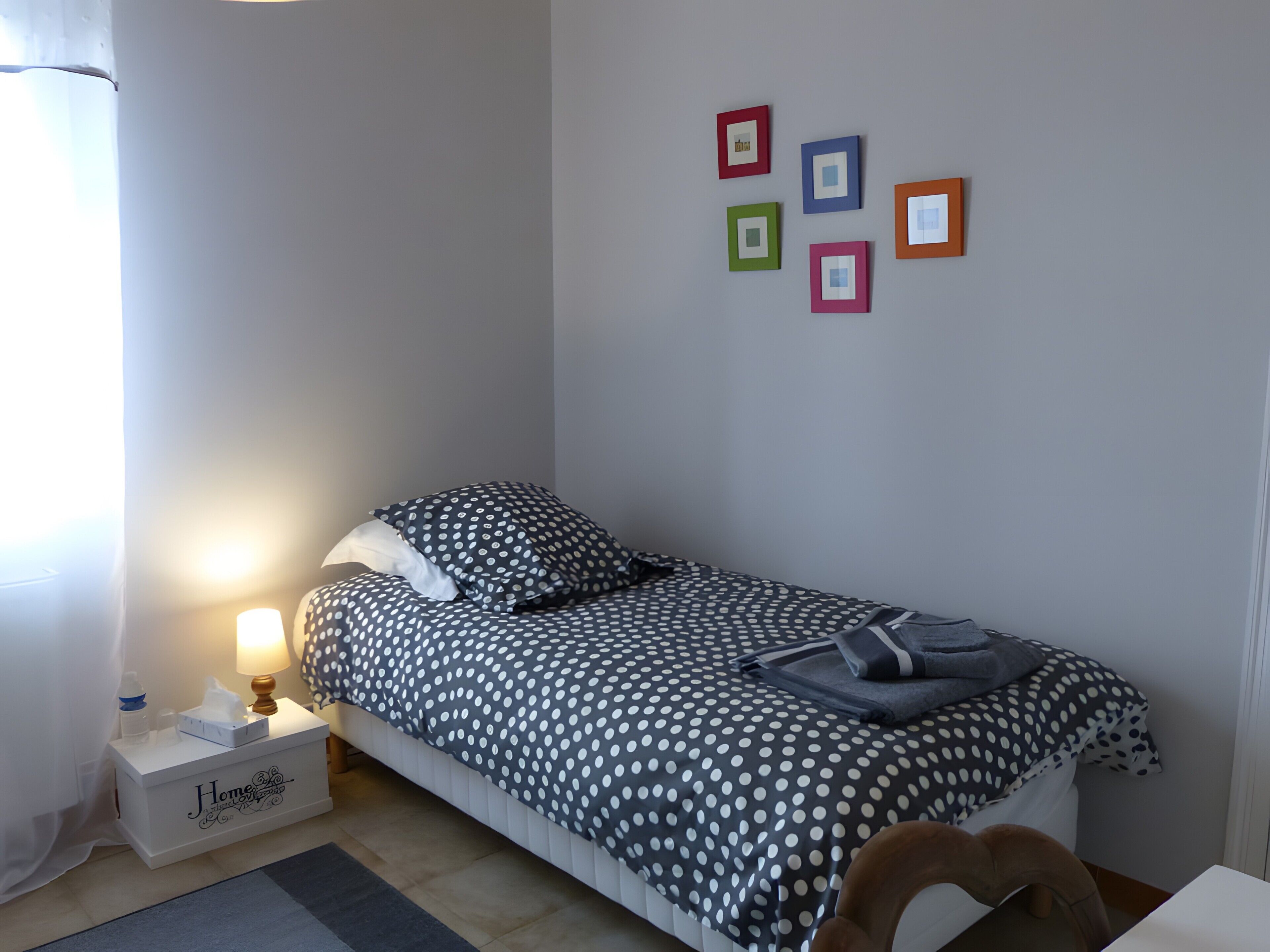Un lit simple avec une couvre-lit à pois, une table de chevet avec une lampe, une petite table de chevet avec une boîte de mouchoirs, et un mur orné de quatre tableaux encadrés.