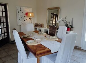 Une salle à manger avec une table en bois dressée pour quatre personnes, des chaises blanches, une lampe, un miroir et un élément décoratif mural.