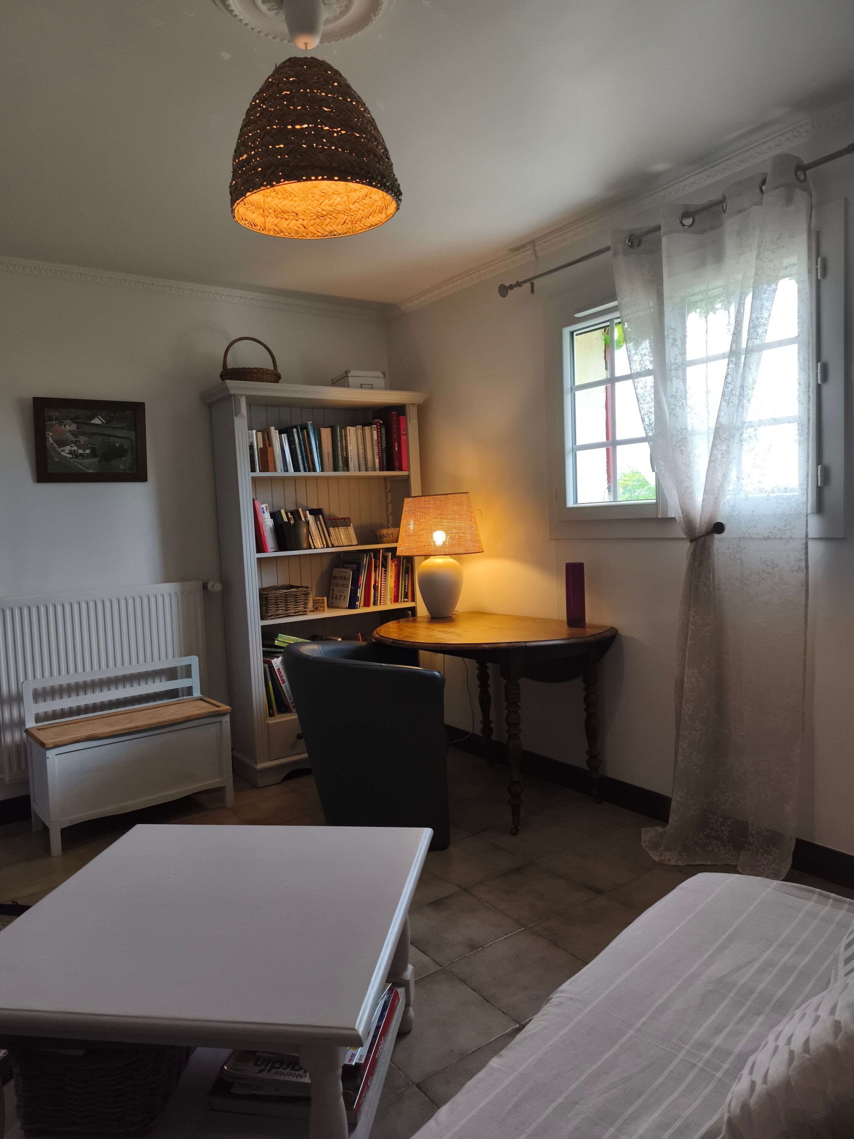 A room with a white desk, a chair, a bookshelf, a small table, a white bench, and a window with curtains.