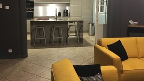 Een moderne woonkamer met gele banken, een keuken op de achtergrond en een zichtbare deur naar een andere kamer.
