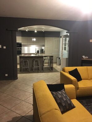 Ein modernes Wohnzimmer mit gelben Sofas, einer sichtbaren Küche im Hintergrund und einer Tür zu einem weiteren Raum.