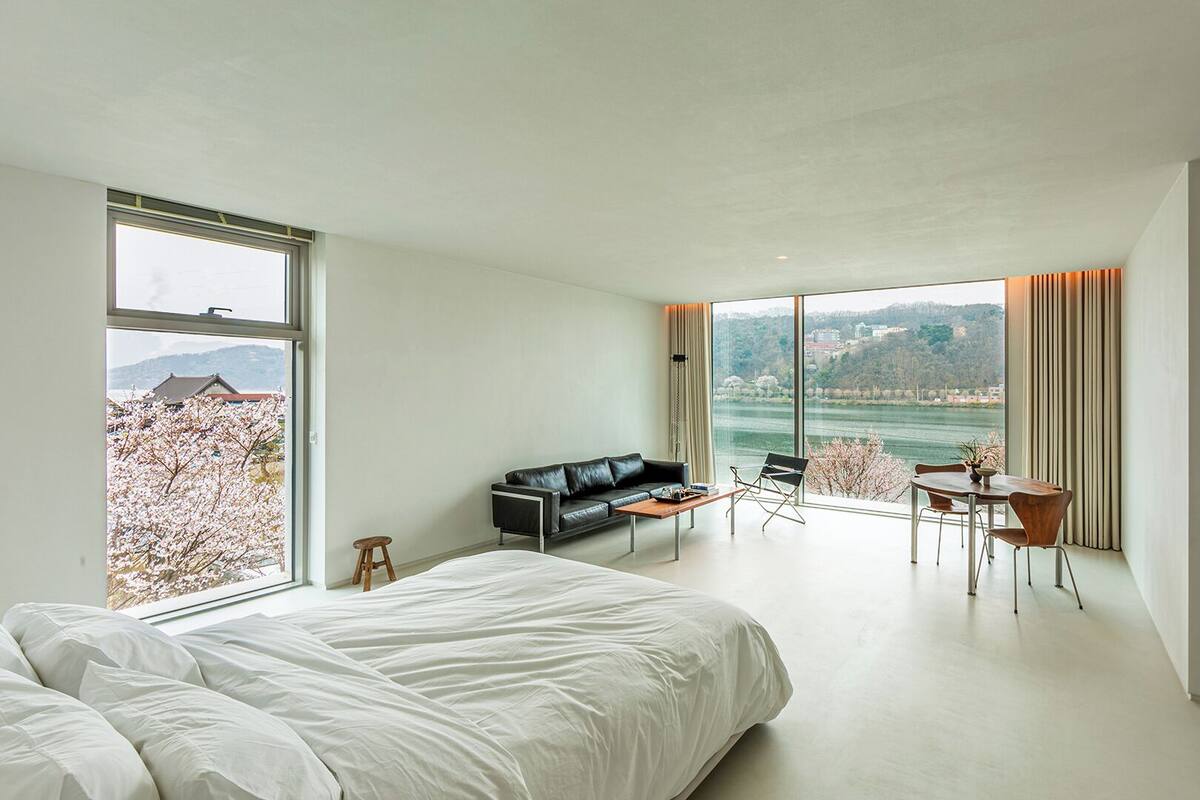 A modern hotel room with a large bed, a sofa, a dining table, and a view of a lake and mountains.
