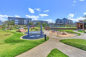 Ein Spielplatz mit Schaukeln und Rutschen, umgeben von grünen Rasenflächen und einem Weg.