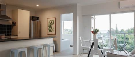 Ein modernes Wohnzimmer mit einer Couch, einem Couchtisch und einem Essbereich mit Stühlen und einem Tisch. Es gibt eine Küchenzeile mit Kühlschrank, Mikrowelle und Spüle. Der Raum verfügt über große Fenster und eine Klimaanlage.