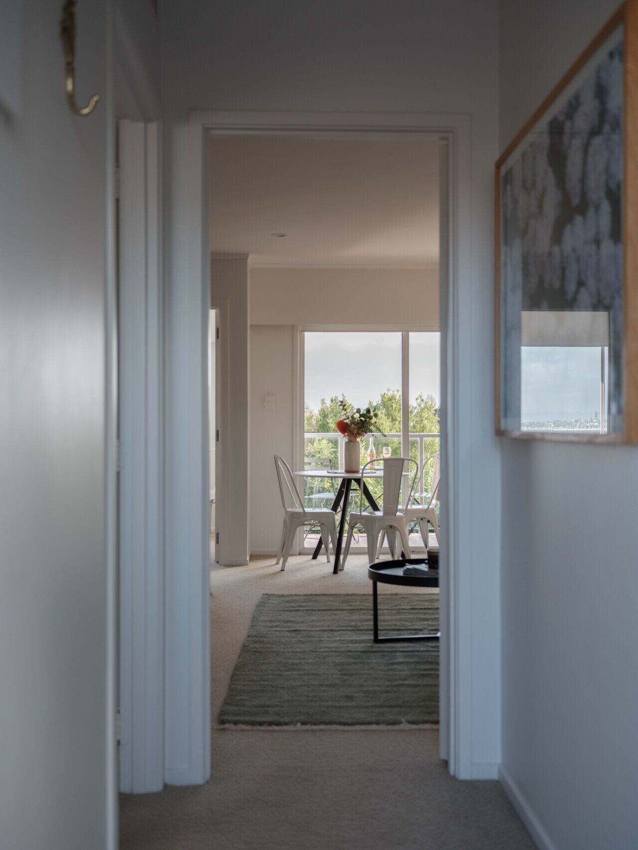 A hallway leading to a balcony with white chairs and a table, a framed picture on the wall, and a view of the cityscape.