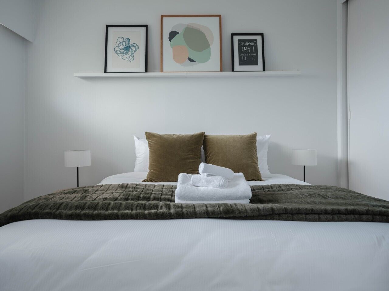 A hotel room with a neatly made bed, two pillows, a blanket, and three framed artworks on the wall.
