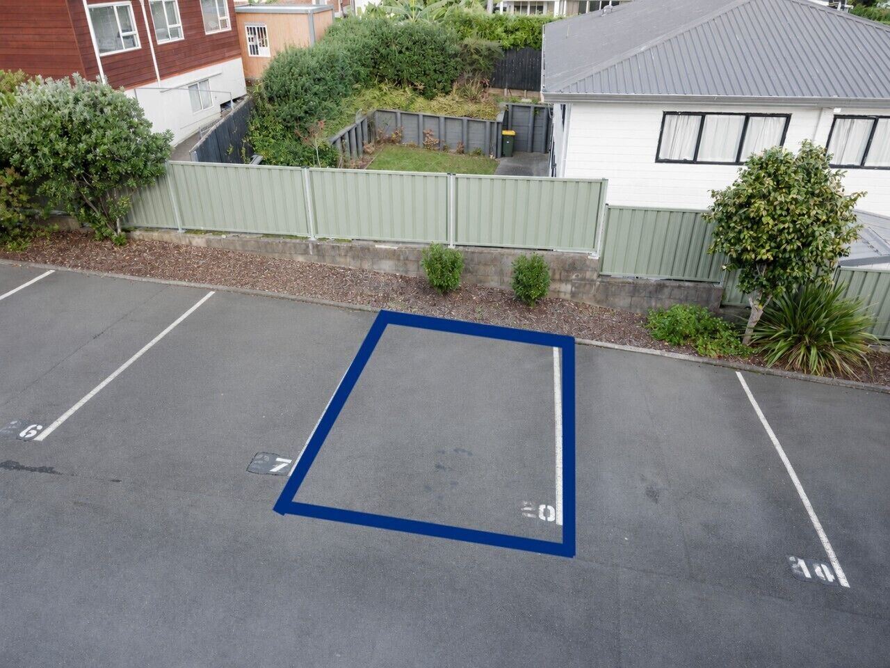 Ein Parkplatz mit markierten Stellplätzen, einem begrenzten Bereich und einem Gebäude mit grauem Dach im Hintergrund.