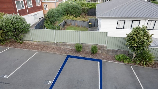 A parking lot with marked spaces, a gated area, and a building with a grey roof in the background. - Walk to Beach | Sky Tower & Ocean Sunset Views | ✨Sage & Sea by Homello✨ (Devonport)