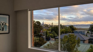 A room with a large window overlooking a cityscape, a framed picture on the wall, and a view of the sky. - Walk to Beach | Sky Tower & Ocean Sunset Views | ✨Sage & Sea by Homello✨ (Devonport)