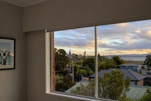 A room with a large window overlooking a cityscape, a framed picture on the wall, and a view of the sky. - Walk to Beach | Sky Tower & Ocean Sunset Views | ✨Sage & Sea by Homello✨ (Devonport)