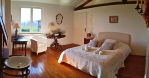 Une chambre avec un grand lit, un bureau avec une lampe, une chaise et une fenêtre donnant sur un paysage verdoyant.