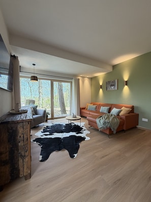 A modern living room with a sofa, a cowhide rug, a wooden table, and a view of trees through large windows.