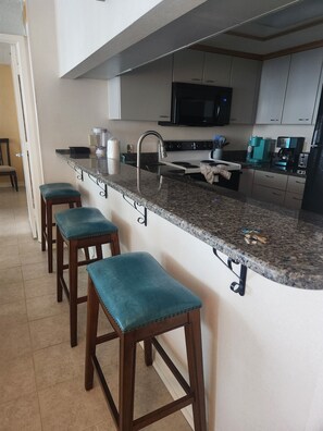A kitchen with a granite countertop, stainless steel appliances, and wooden stools. - Majestic neon sunsets from the balcony of this oceanview 2bdrm 2bth condo. (Myrtle Beach)