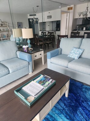 A living room with two light blue sofas, a coffee table with magazines, a dining area with chairs, and a kitchen in the background. - Majestic neon sunsets from the balcony of this oceanview 2bdrm 2bth condo. (Myrtle Beach)