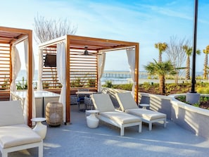 Un salon sur le toit doté de pergolas en bois, de fauteuils confortables avec des coussins blancs, d’une petite table et d’un ventilateur de plafond.