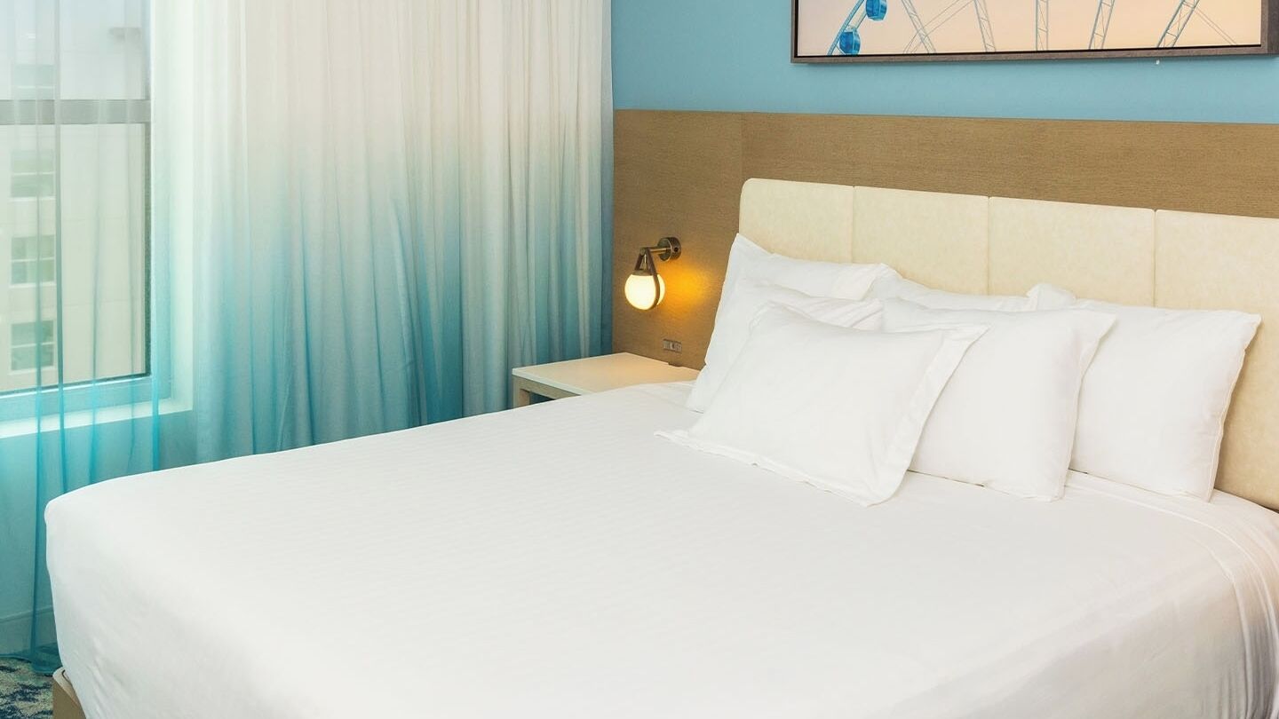 A hotel room with a large bed, a nightstand with a lamp, a window with sheer curtains, and a wall-mounted artwork.