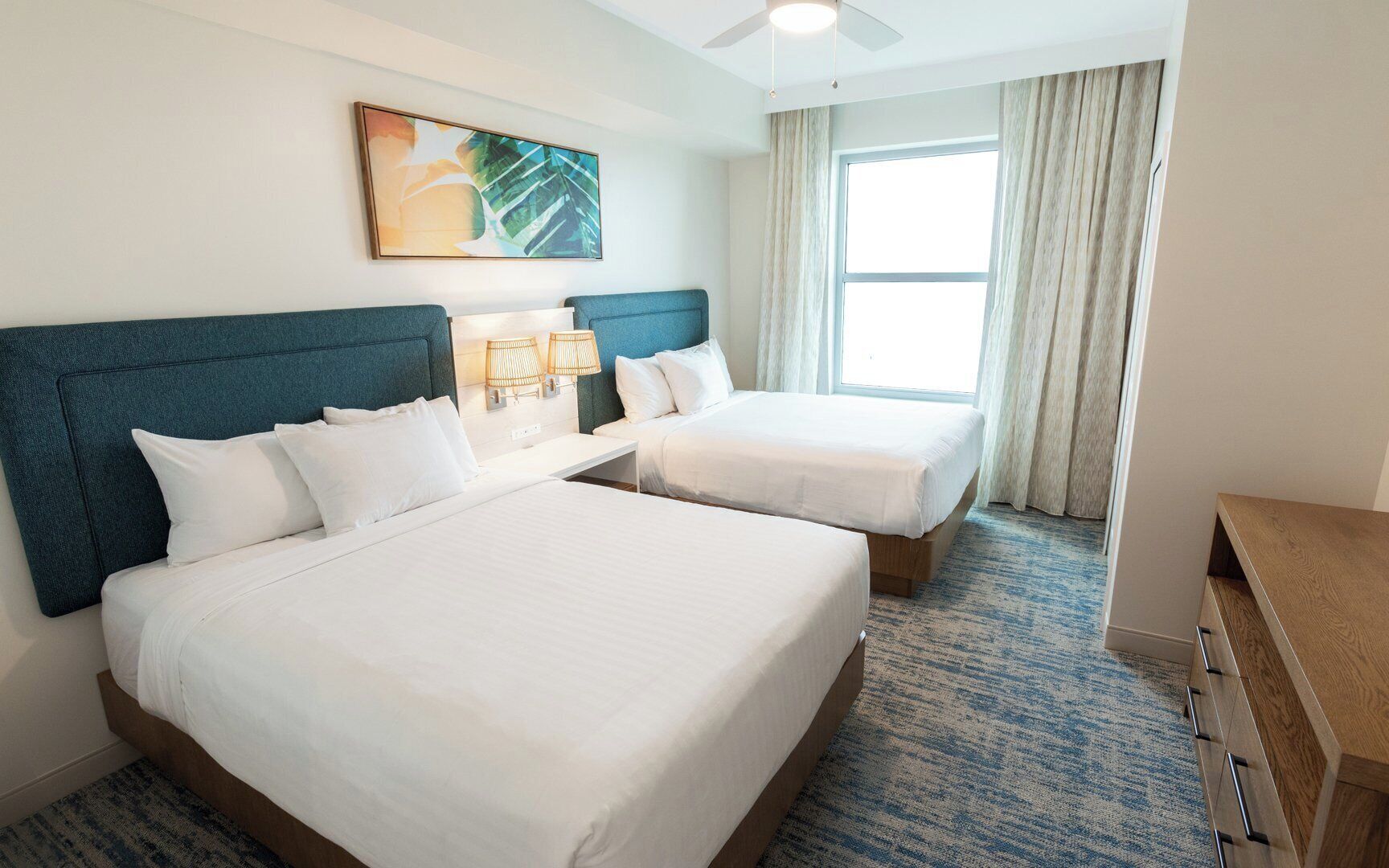 A hotel room with two beds, a ceiling fan, a painting, and a large window with curtains.
