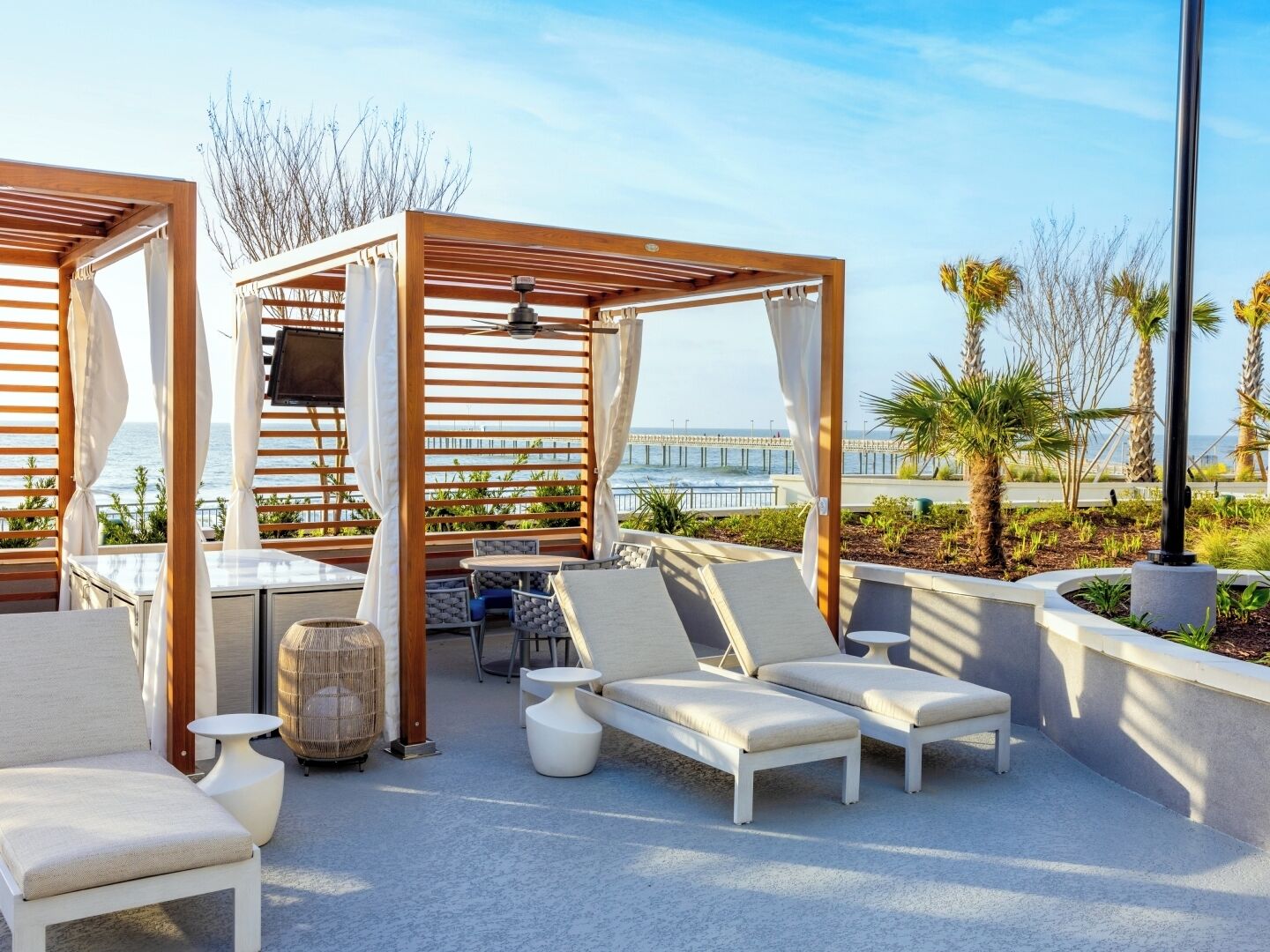 A rooftop lounge with wooden pergolas, white cushioned lounge chairs, a small table, and a ceiling fan.