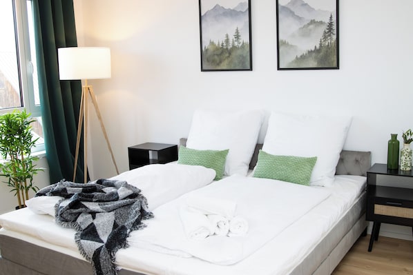 A neatly made double bed with white linens, a green throw blanket, and a rolled towel. There are two green pillows, a bedside table with a lamp, and a framed picture of a mountain landscape on the wall.