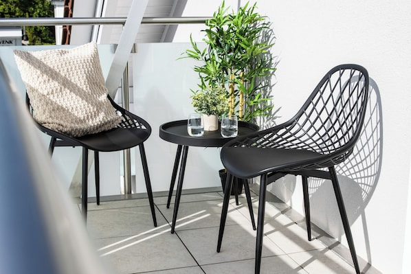 Deux chaises noires avec un coussin à motifs tissés, une petite table avec deux verres d’eau et une plante en pot sur un balcon.