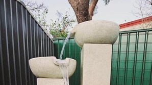 Une fontaine en pierre avec de l’eau en cascade, placée devant une clôture verte et un arbre.