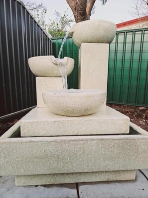Une fontaine en pierre avec de l’eau en cascade, placée devant une clôture verte et un arbre.